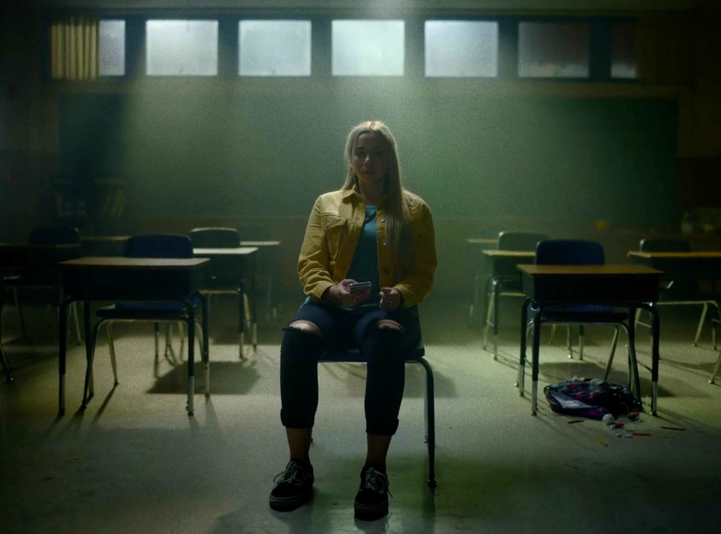 A cinematic wide shot of a woman sitting alone at a desk in a dark, moody room.