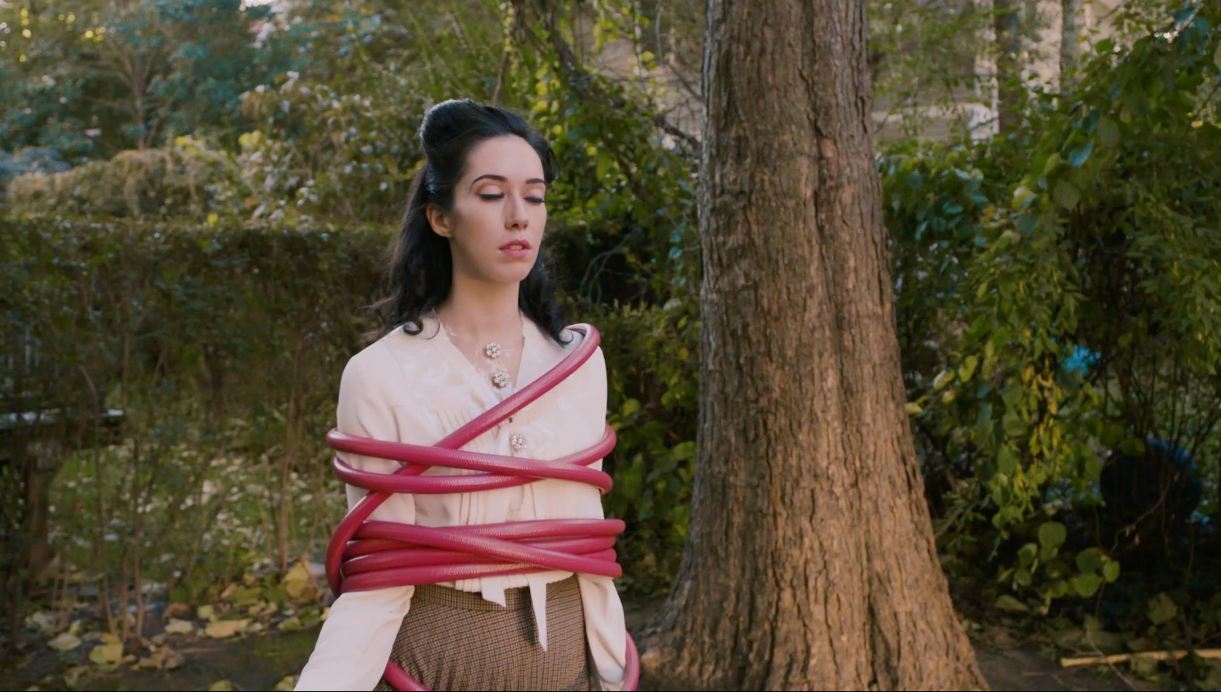 Production still showing a woman with eyes closed, bound in a pink hose, demonstrating conceptual art direction and lighting in an outdoor natural location for creative film production.