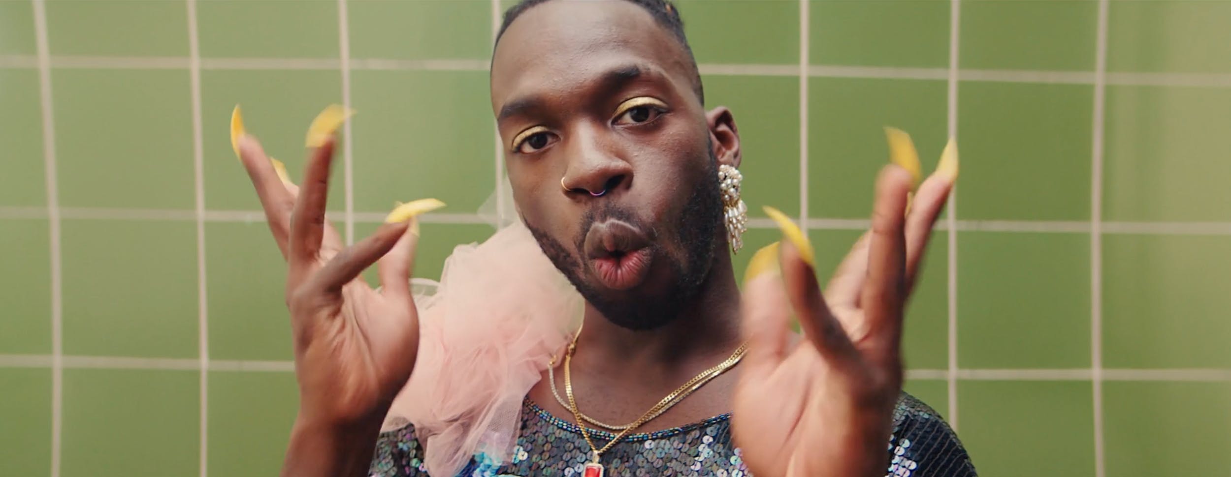 Close-up of a performer with long yellow nails and sequins against green tiles. Bold art direction and stylized lighting for a high-fashion editorial or queer-centric digital series.