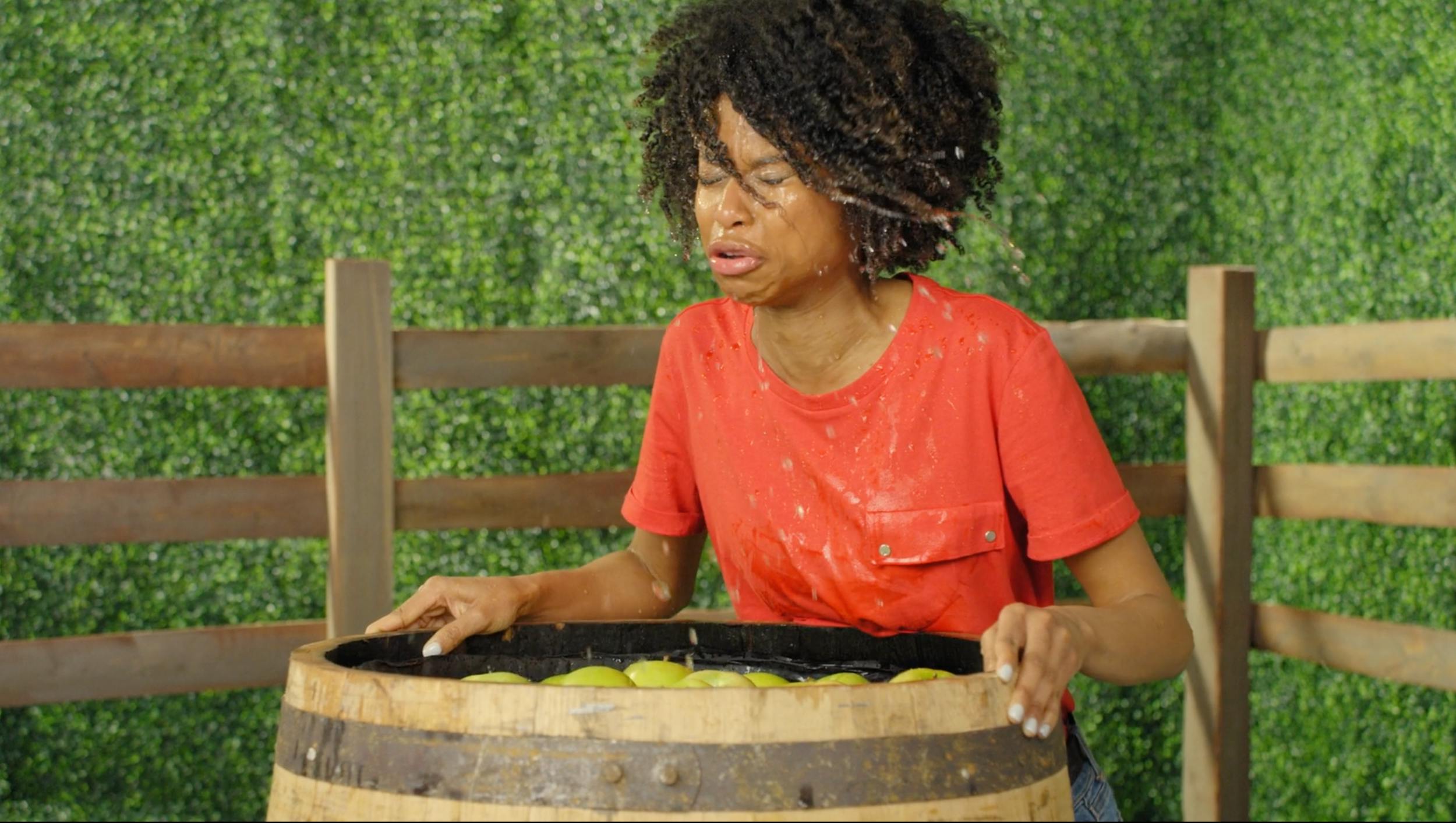 Action shot of a woman getting splashed while apple bobbing. High shutter speed captures water droplets in mid-air. Vibrant color palette for energetic, youth-oriented commercial content.