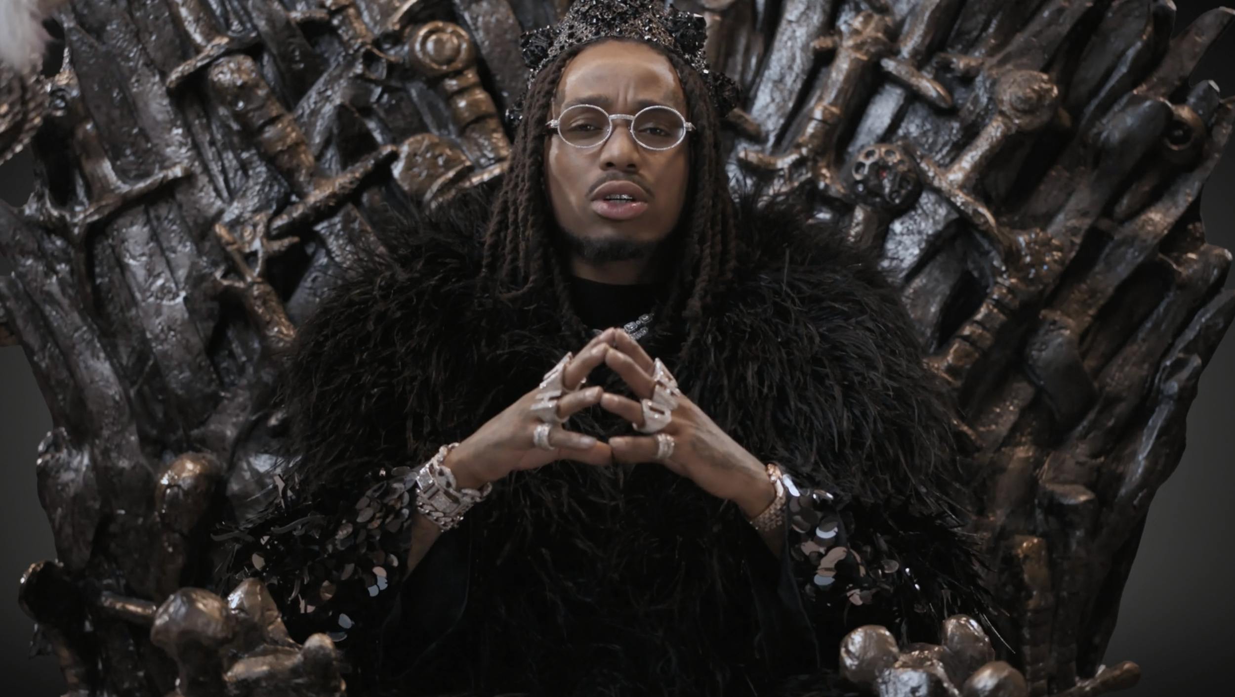 A man with glasses and jewelry sitting on an ornate, 