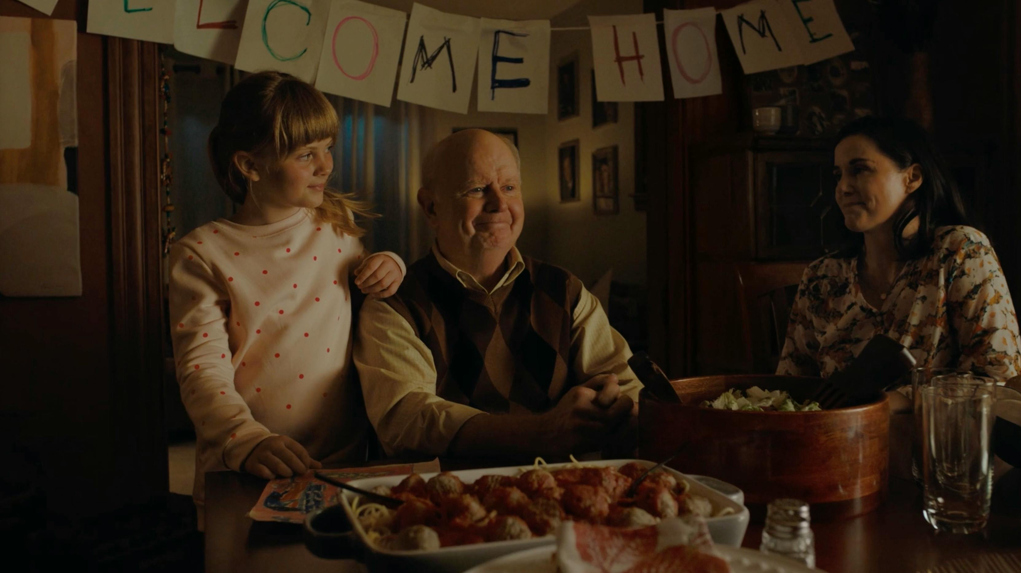 A warm, domestic scene featuring an older man and a young girl at a dinner table. The lighting is warm and motivated, appearing to come from an overhead dining fixture, creating a cozy, 