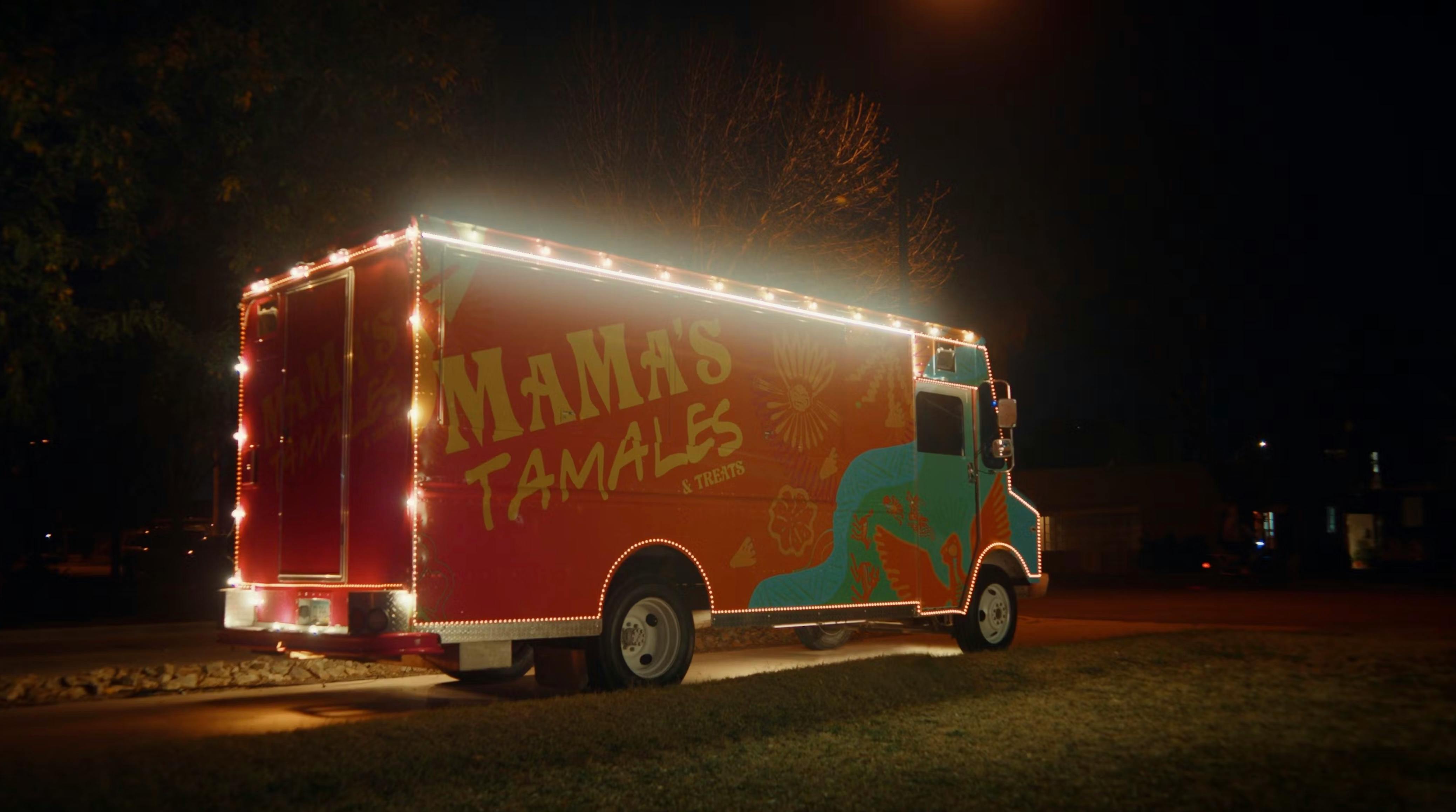 Wide shot of a vibrant orange food truck at night, illuminated by a string of warm marquee lights along the roofline. The truck features stylized 