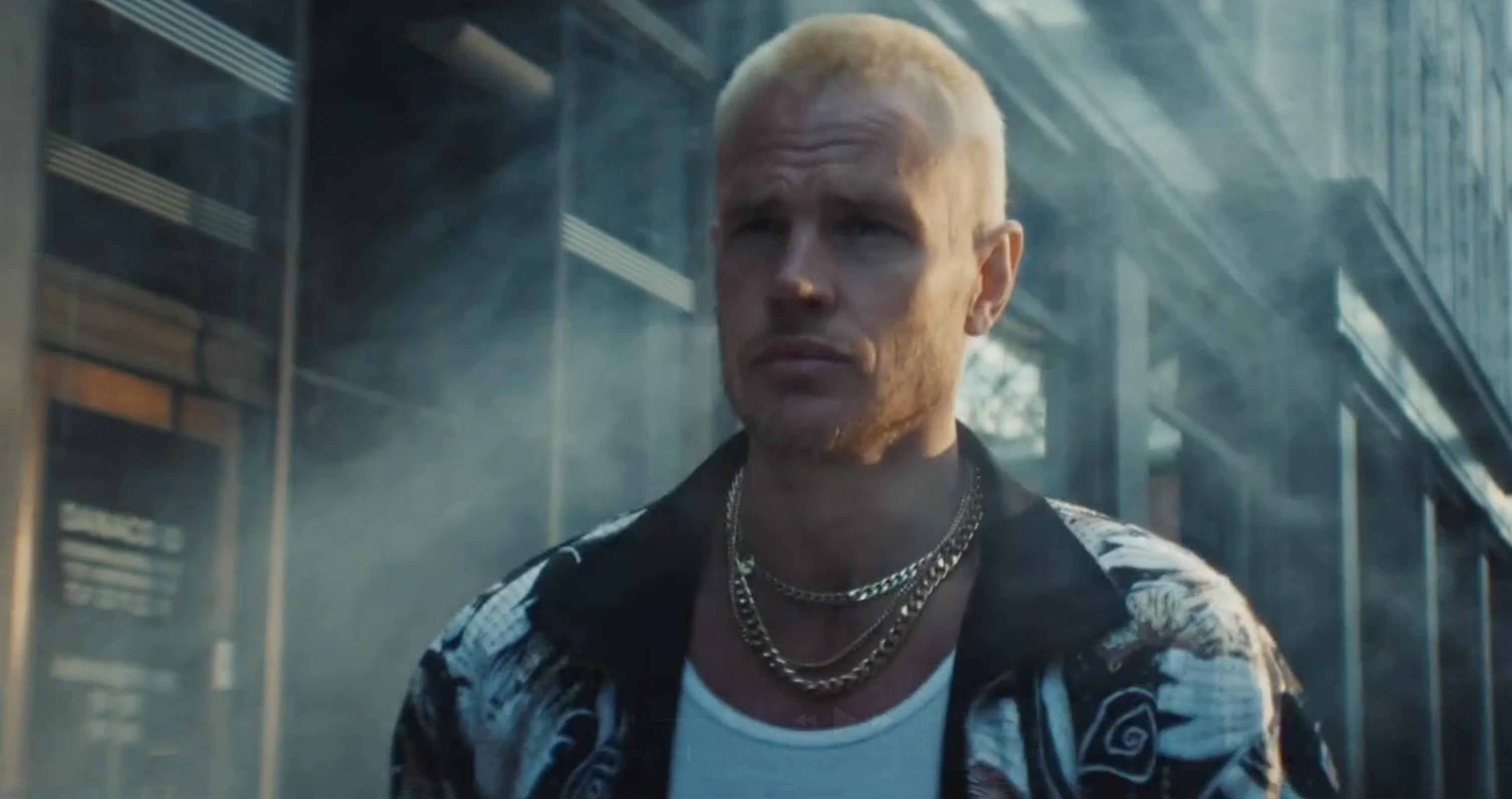 A man with blonde hair and a necklace in a stylized, blue-toned shot.