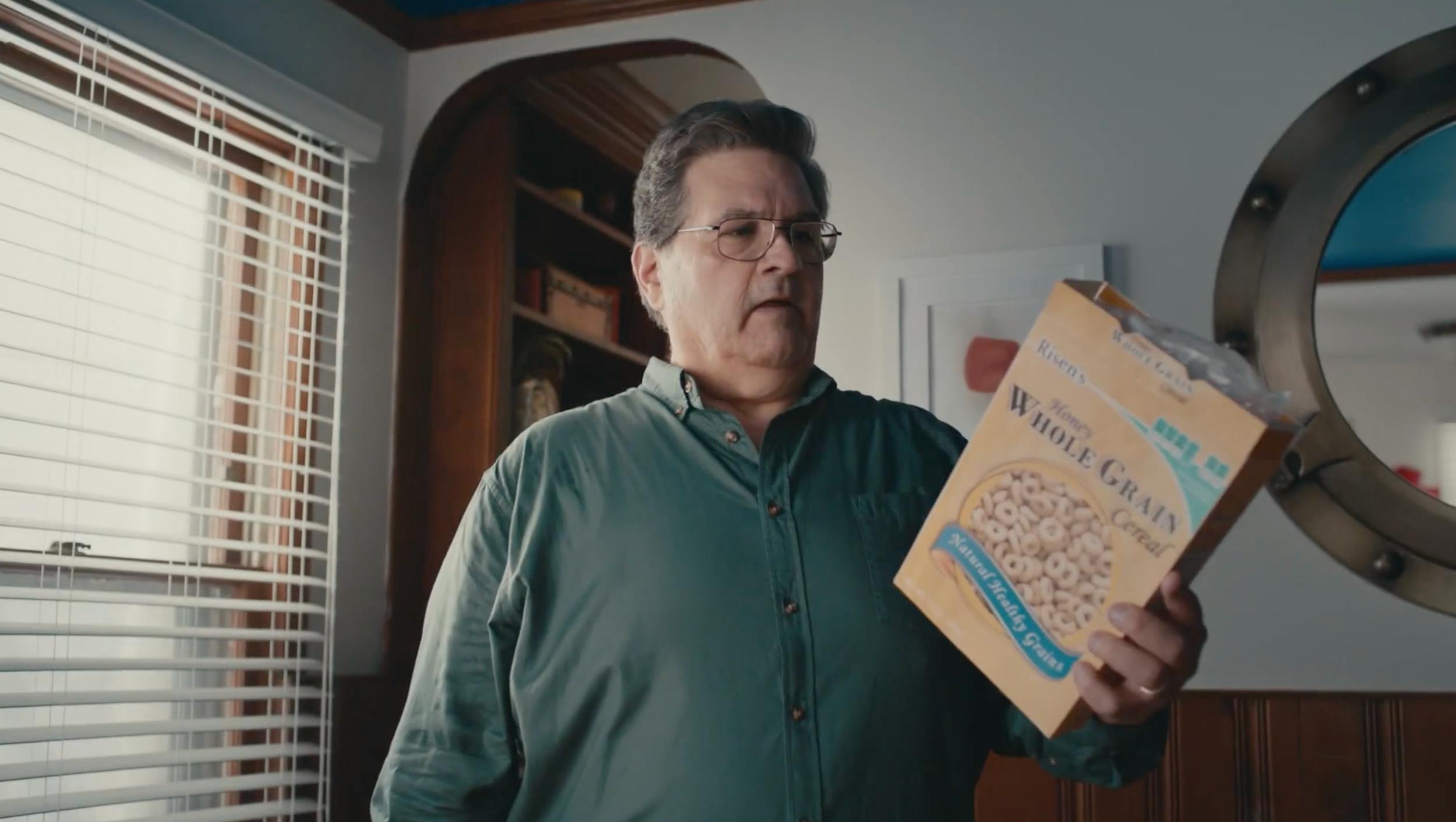 : Interior medium shot of a man in a green shirt holding a cereal box. Naturalistic lighting and shallow depth of field create a domestic, authentic aesthetic for product placement scene