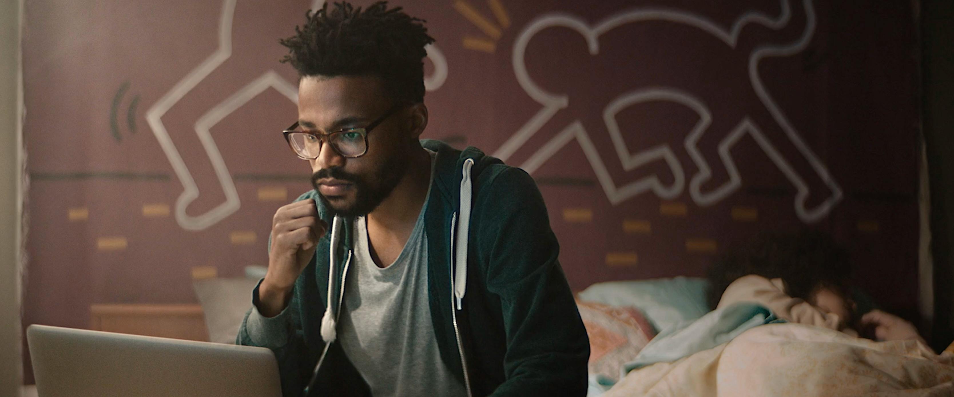 Close-up of a man with glasses and a beard, looking intently at a screen. The lighting is moody and directional, casting soft shadows on his face, with a background featuring graphic wall art and soft interior bokeh.