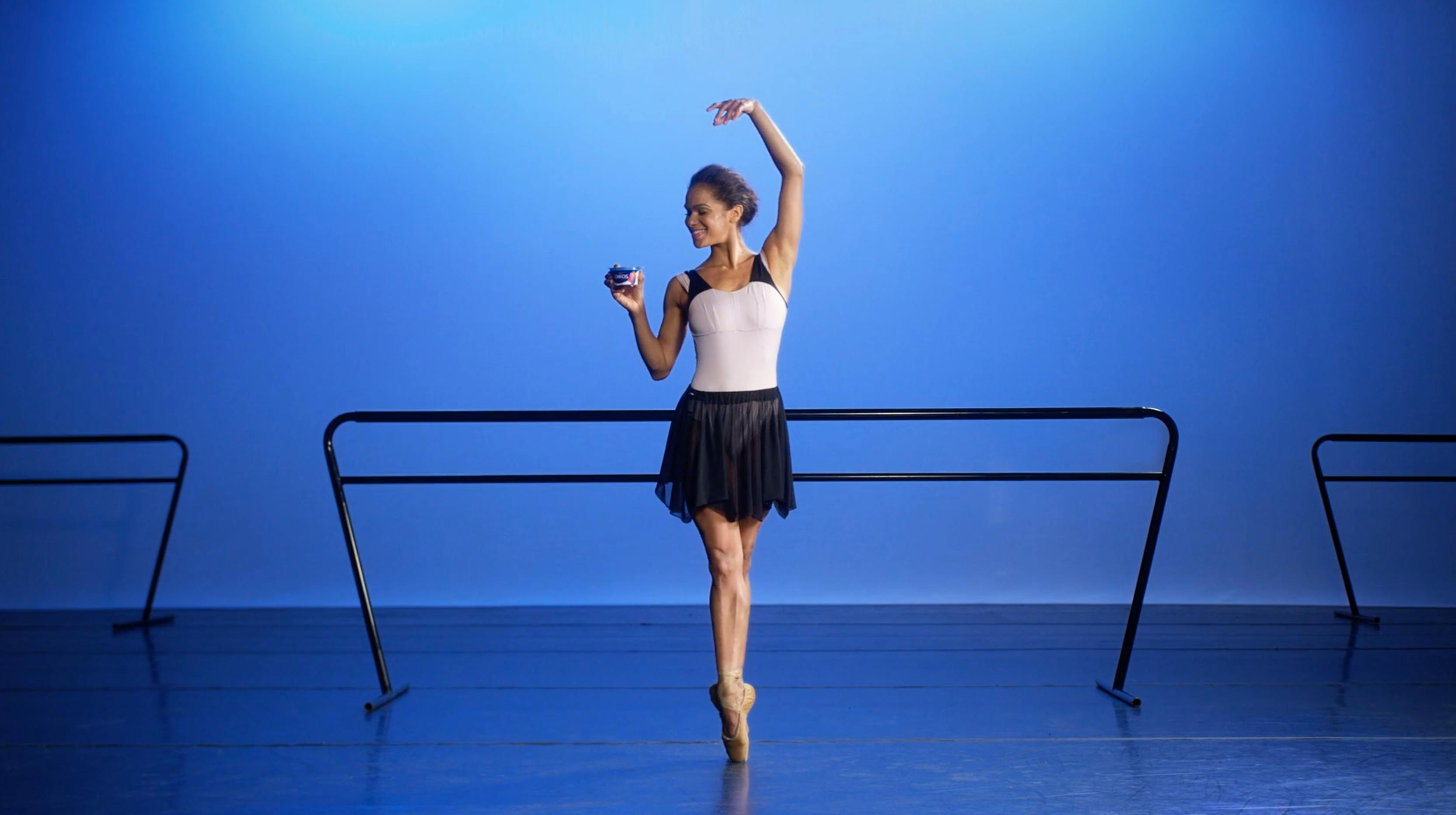 Full shot of a ballerina in a white leotard and black tutu practicing at a barre. The background is a solid cerulean blue cyc wall, evenly lit with high-key studio lighting to emphasize her silhouette and form.