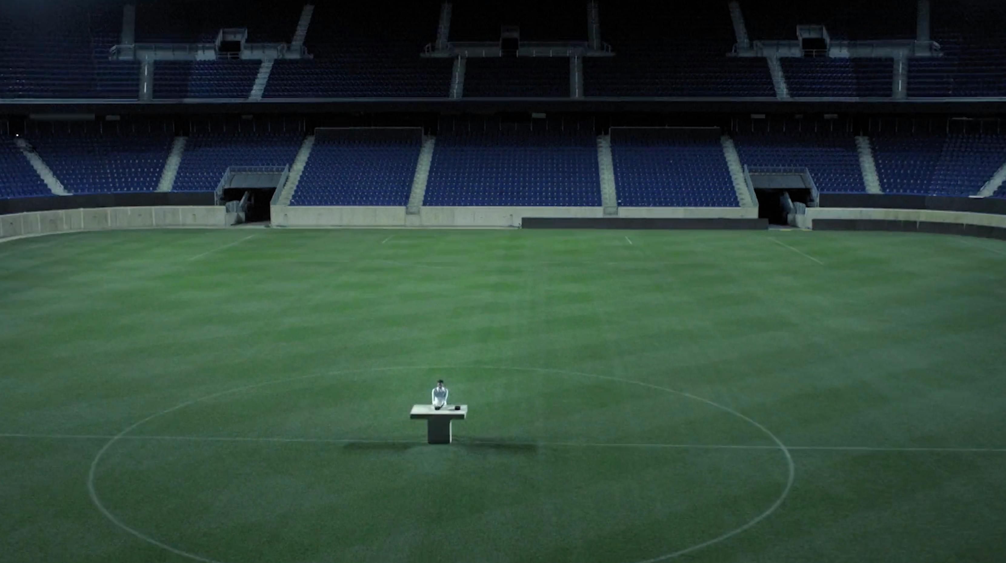 Wide establishing shot of a lush green sports field at night. The empty bleachers are shrouded in shadow, while the turf is illuminated by overhead stadium floodlights, creating a high-contrast, symmetrical composition.