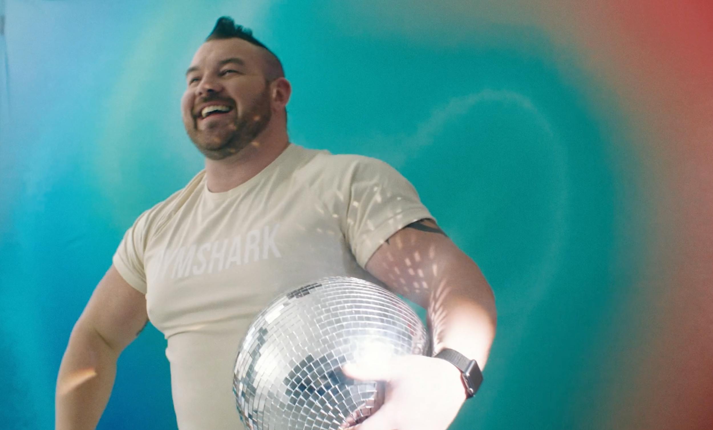 A smiling, muscular man with a mohawk haircut holding a large disco ball. He is wearing a branded 