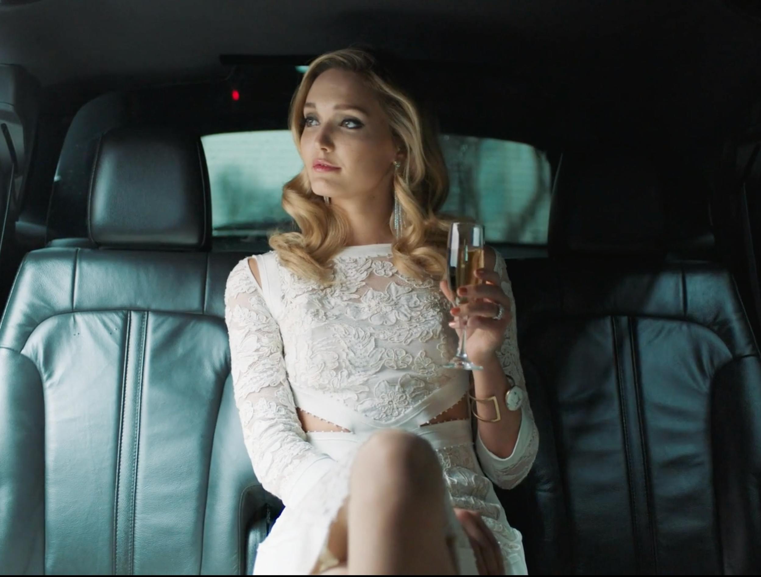 A sophisticated blonde woman in a white lace dress sitting in the back of a luxury vehicle, holding a glass of champagne. Soft side-lighting emphasizes her profile against the dark car interior.