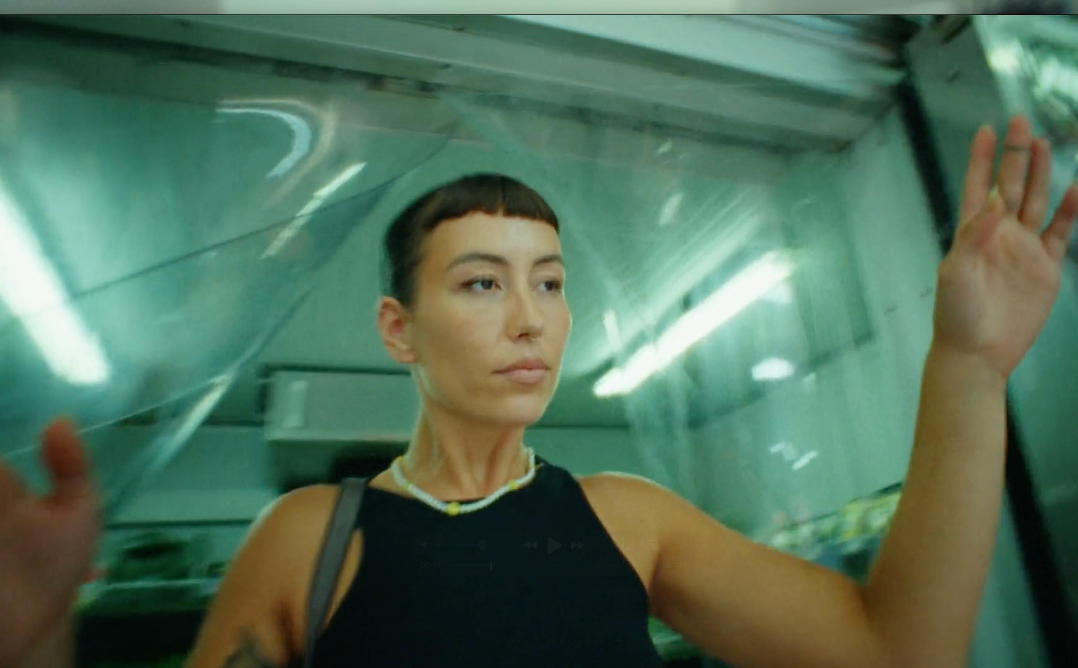 A person with short hair and tattoos on their arm sits in a modern interior with cool, greenish lighting, looking thoughtfully off-camera.