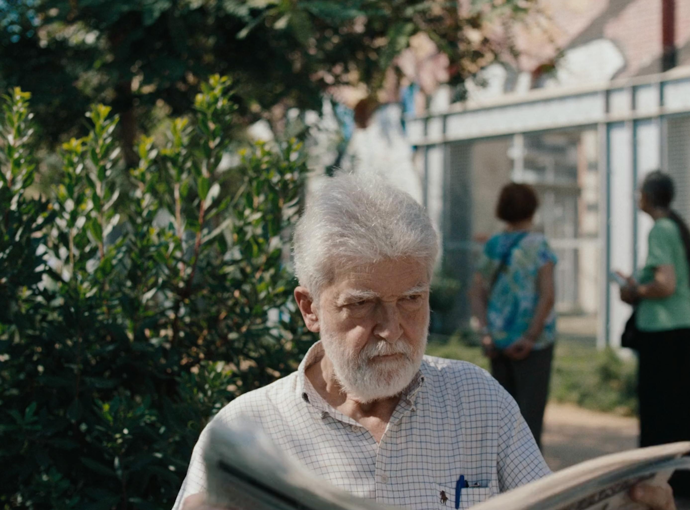 An older man with white hair and glasses looks down at something in his hands while standing in an outdoor, sunlit courtyard.