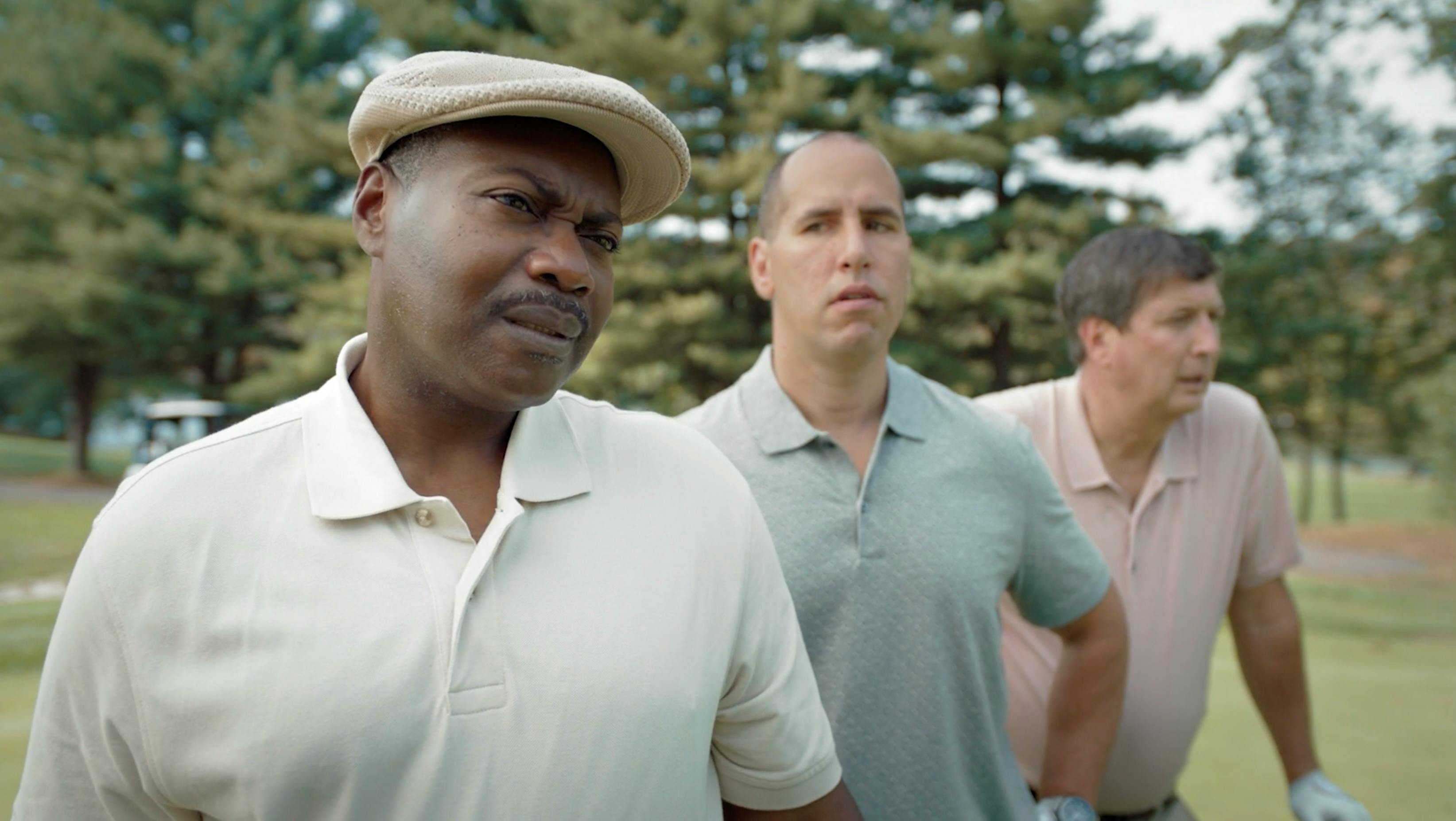Medium shot of two men outdoors in natural, diffused daylight. The foreground subject wears a white polo and tan flat cap, captured with a shallow depth of field that softly blurs the green foliage in the background.