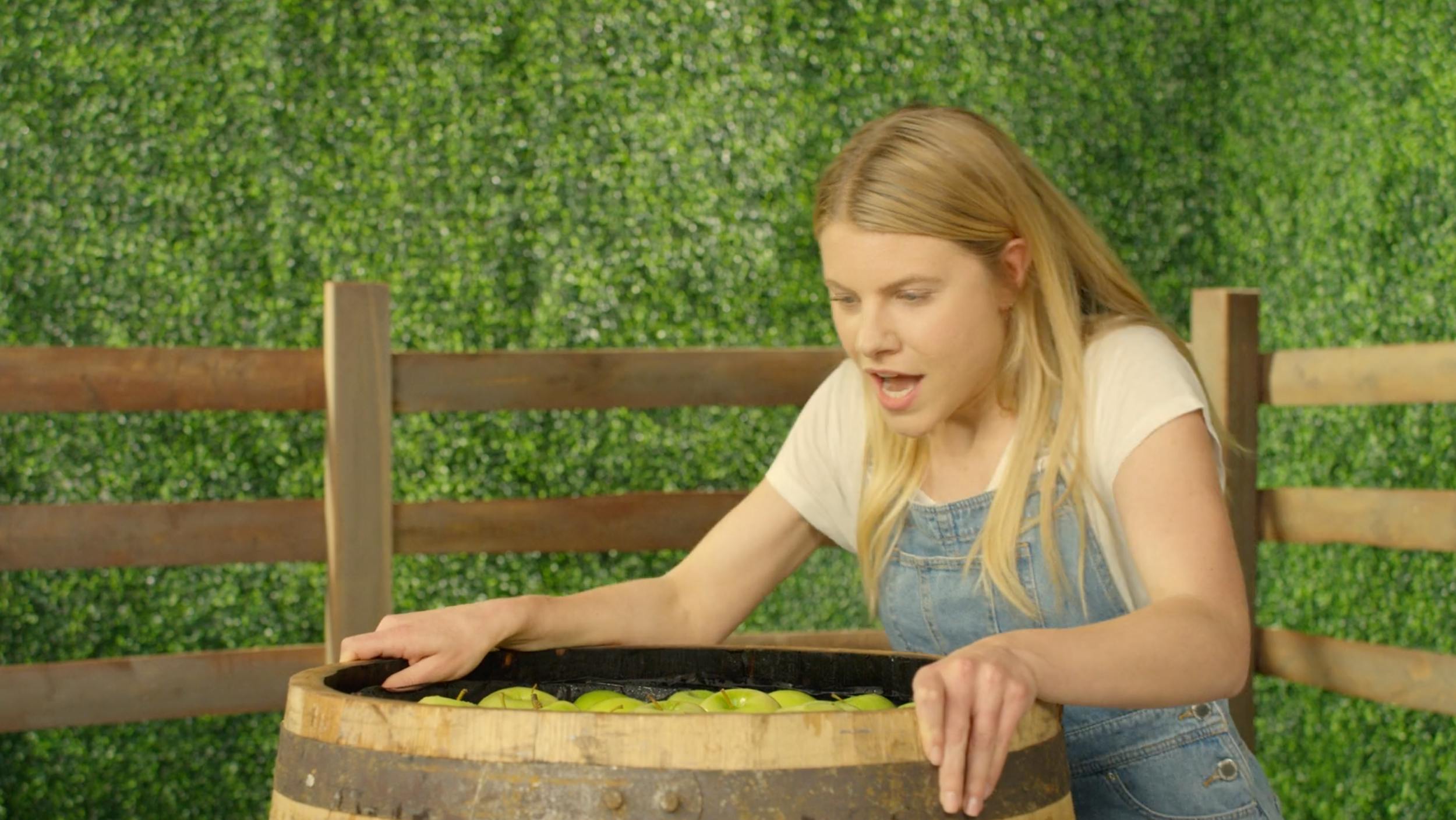 Medium shot of a female actor bobbing for apples in a rustic wooden barrel. Bright, even lighting and a green hedge backdrop suitable for lifestyle promos and high-frame-rate b-roll.
