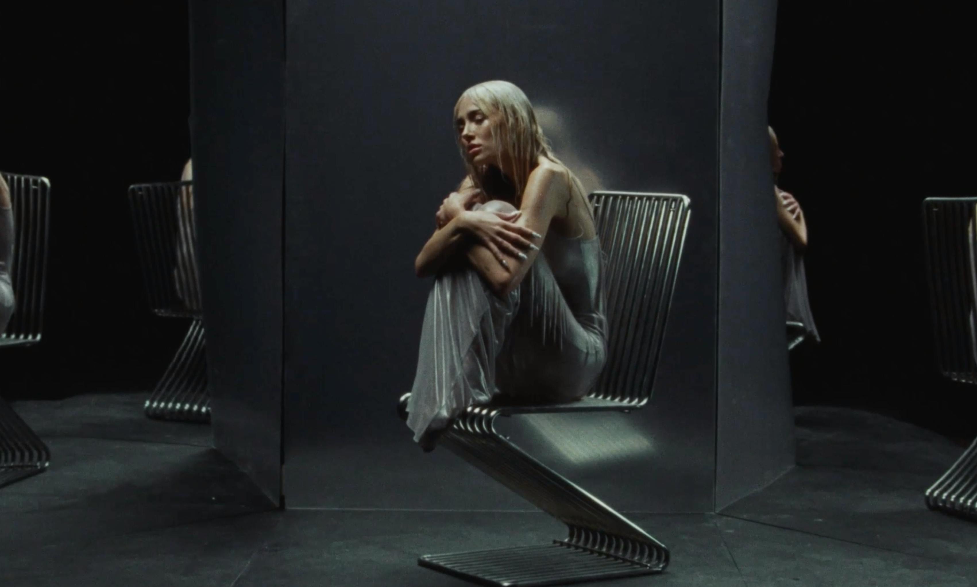 A woman in a dress sits alone in a dark, minimalist studio space with a single spotlight hitting her.