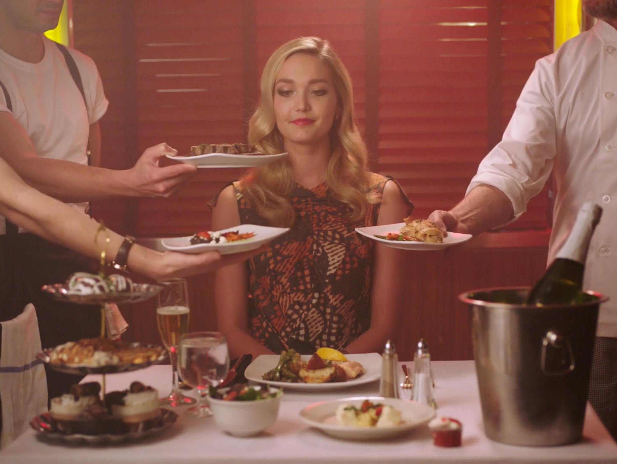 A blonde woman in an elegant dress seated at a lavish dinner table while servers present multiple gourmet plates. The scene features warm, moody red-toned lighting and a shallow depth of field for a high-end luxury vibe.