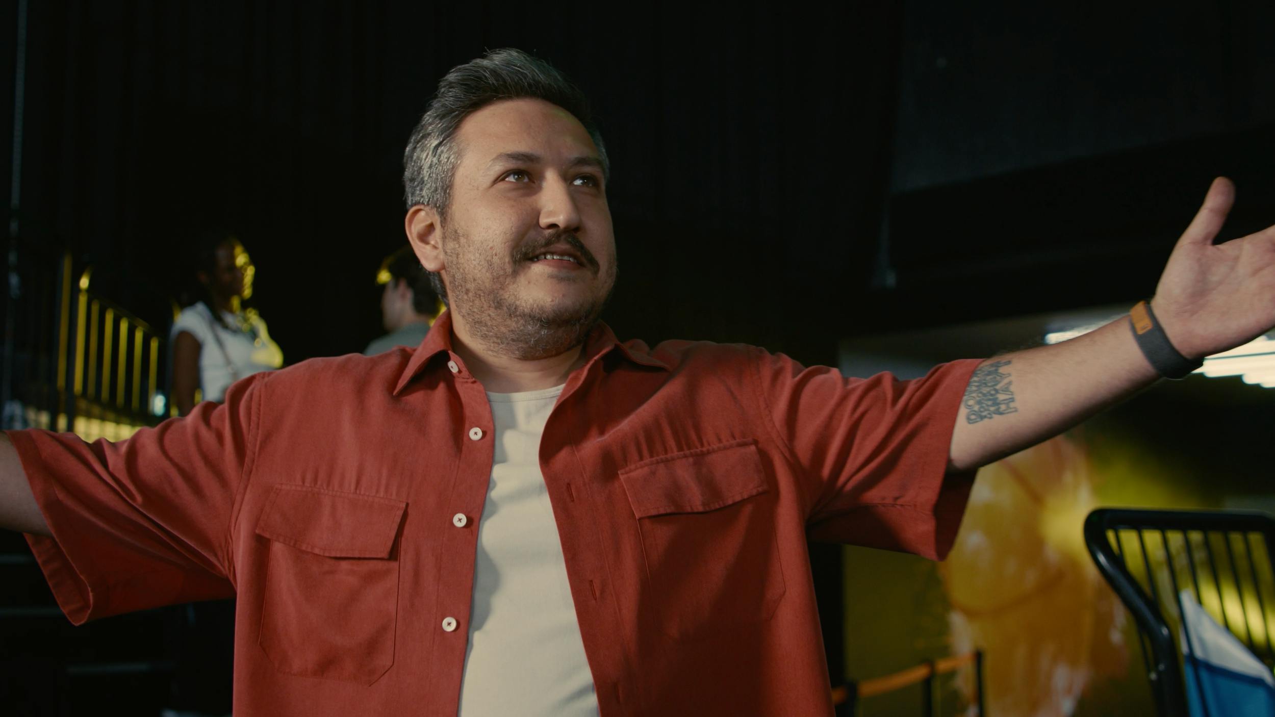 Confident man in a red shirt presenting with open arms in a vibrant indoor setting, capturing personality-driven storytelling in commercial video production.