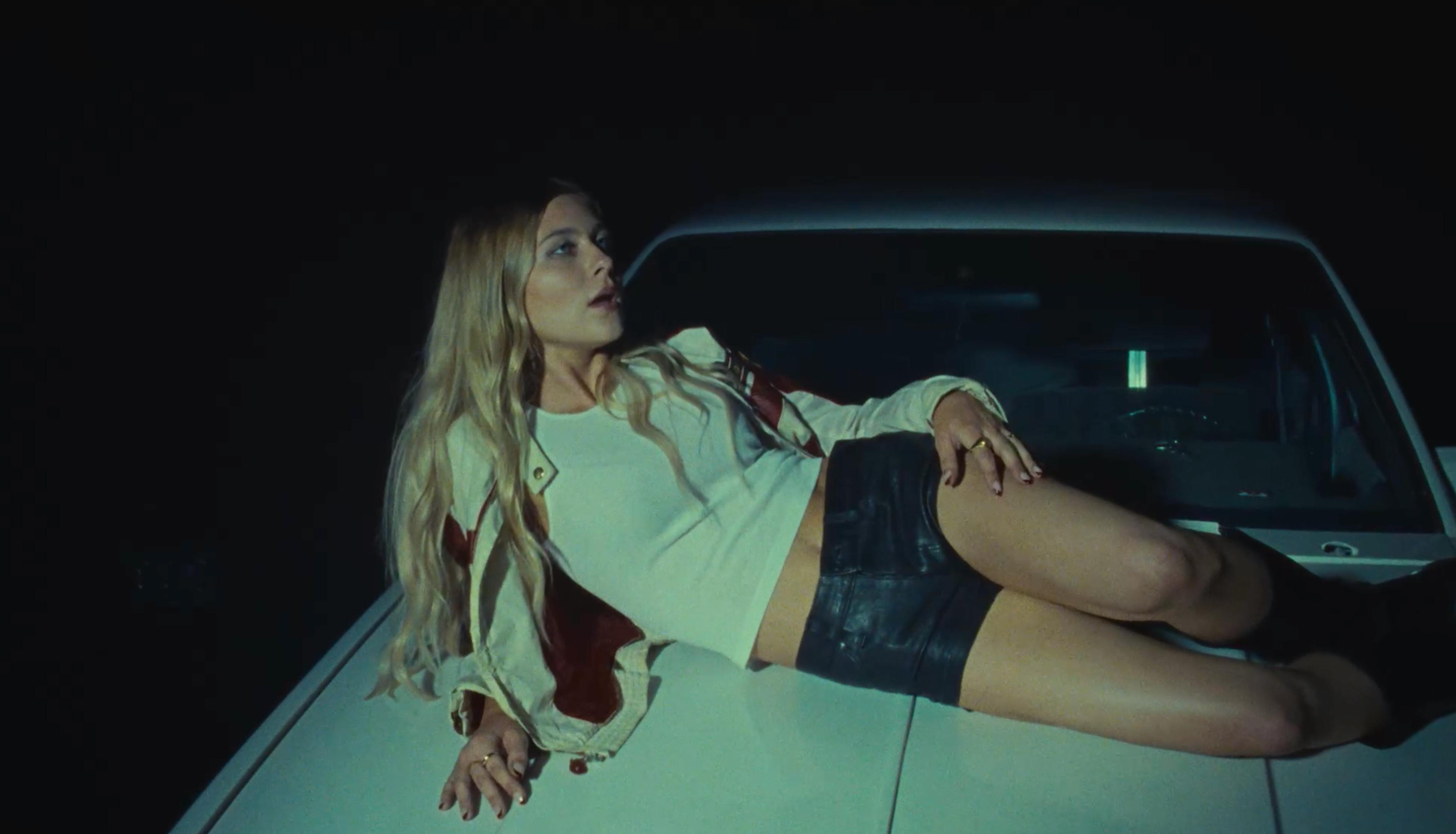A woman with blonde hair lies on a dark surface, leaning on her elbow and looking toward the camera in a moody, low-lit environment.