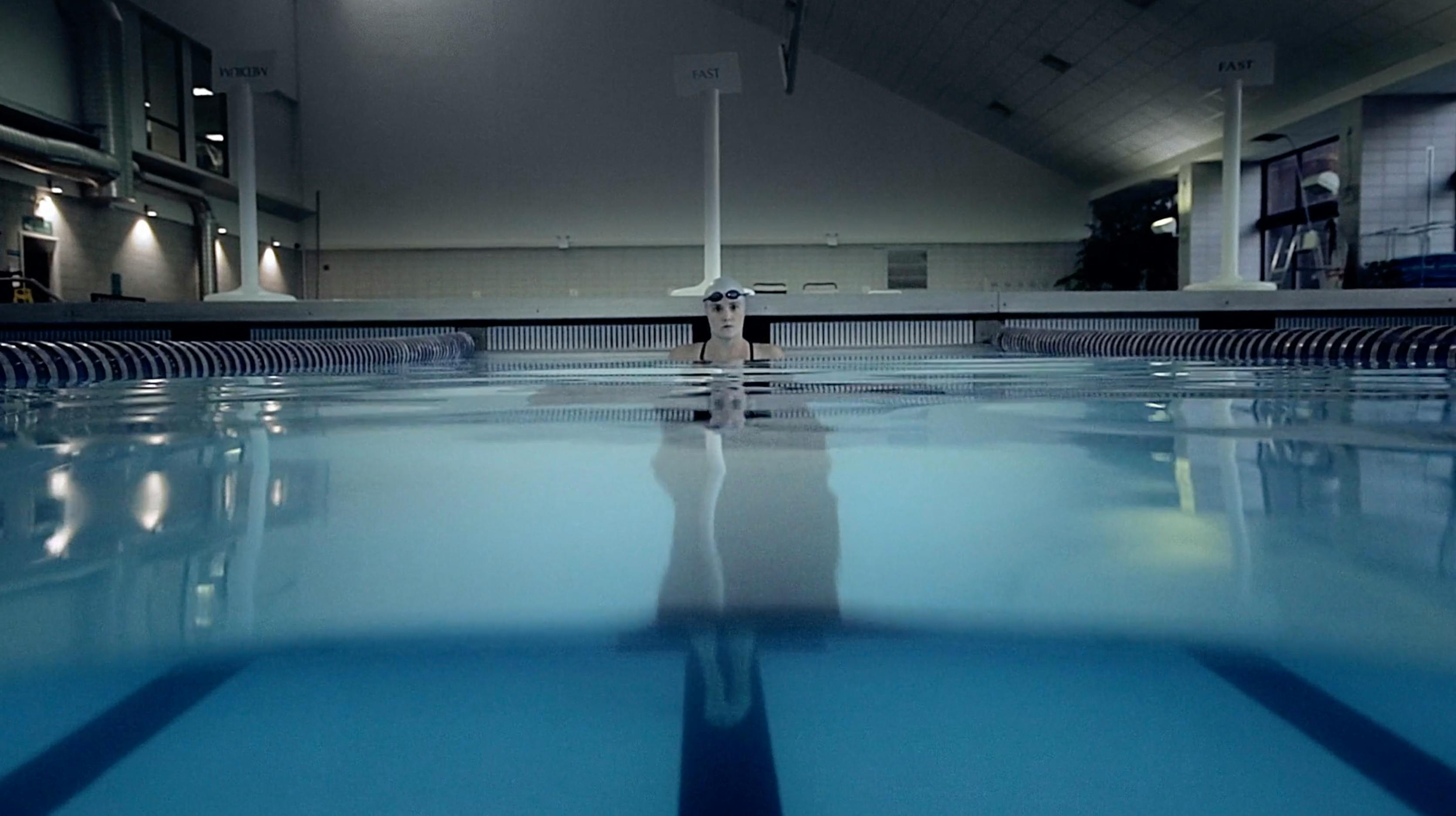 A cinematic low-angle shot of an indoor swimming pool with calm, reflective blue water. A diving board stands at the far end under soft, moody overhead lighting, creating a serene and symmetrical architectural composition.