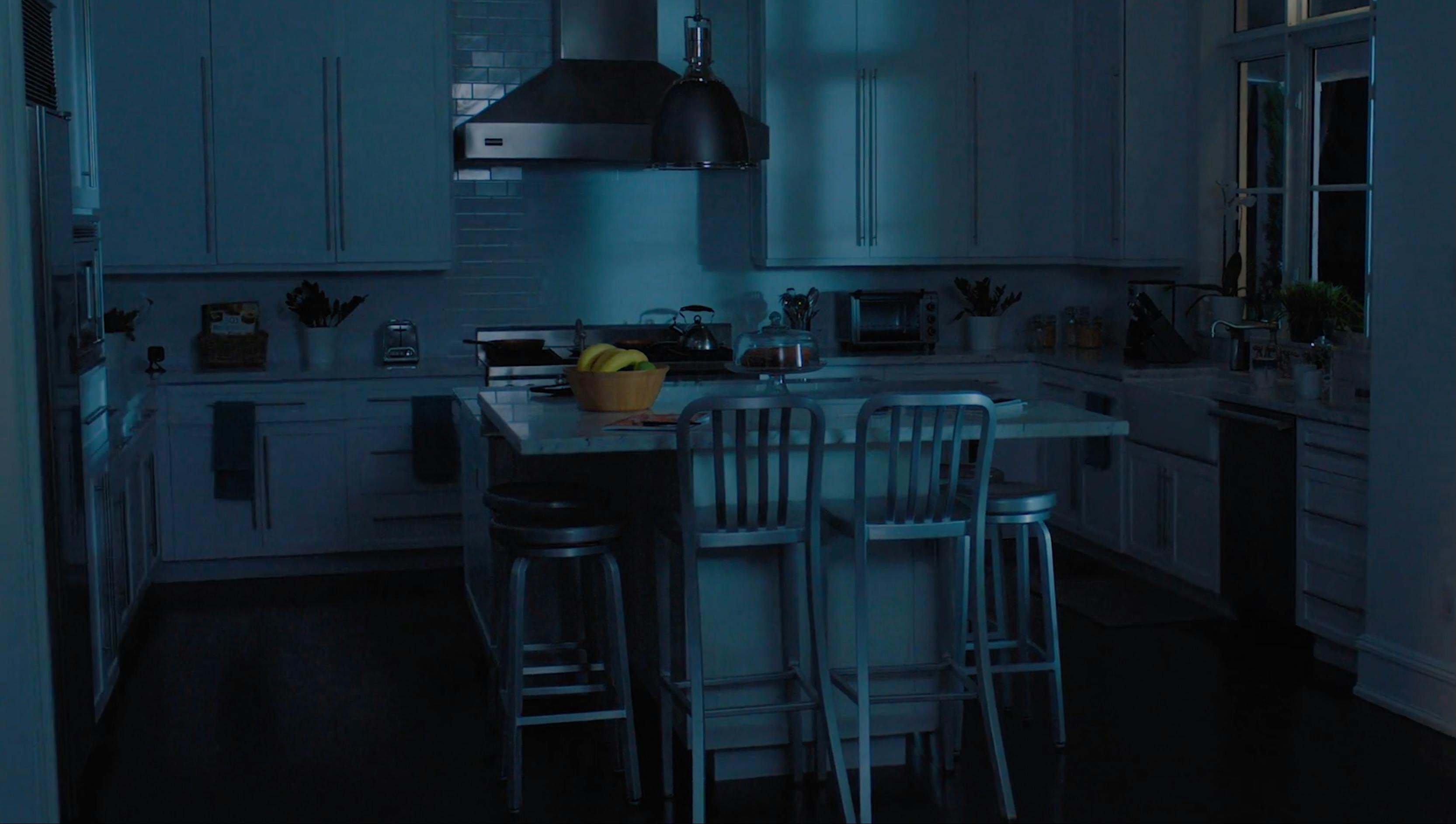 A wide shot of a modern kitchen island with blue cabinetry and wooden bar stools. The scene is lit with cool, nighttime ambient light, showcasing professional interior set design and cinematography.