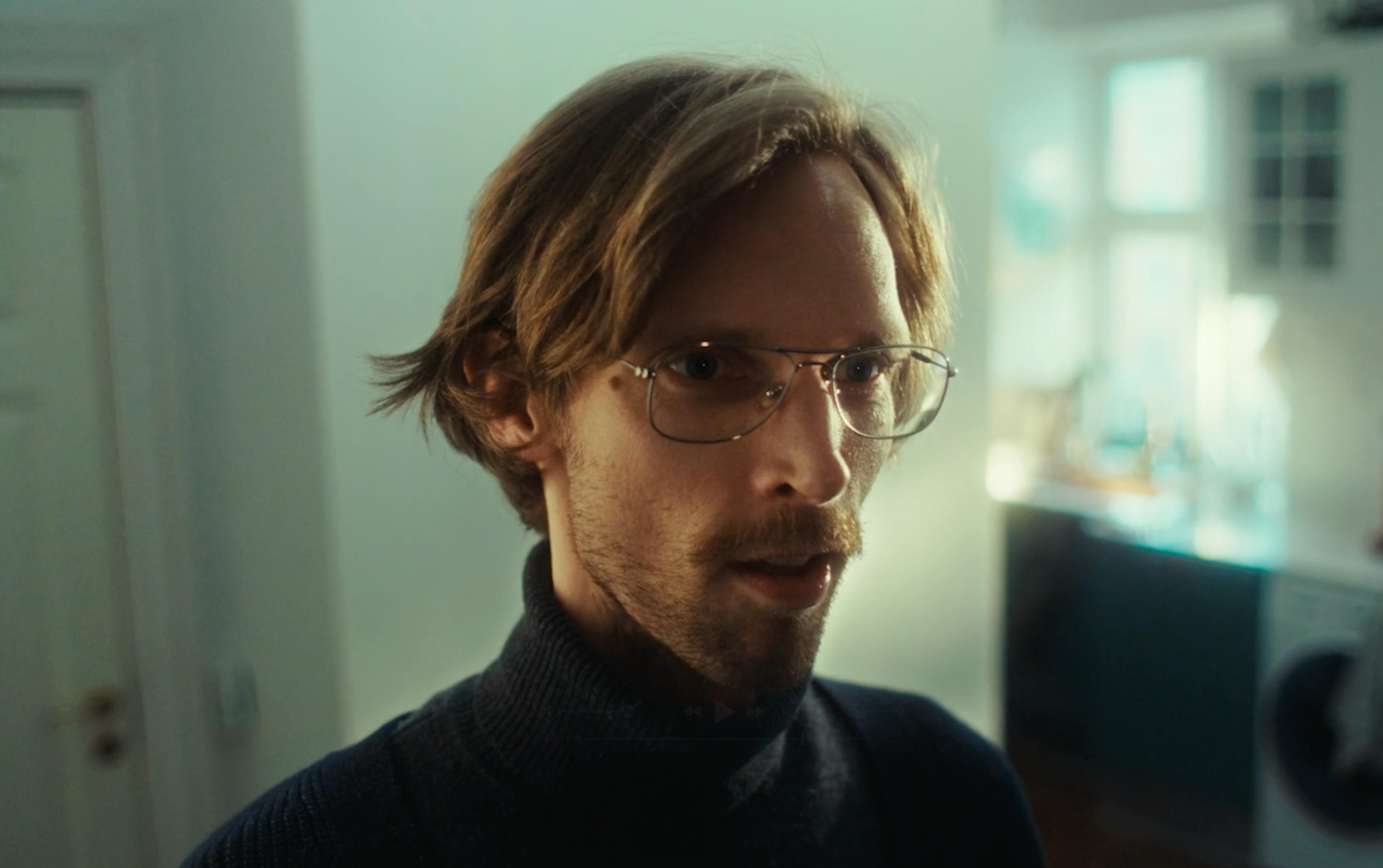 A medium close-up of a man with light brown hair and a goatee, wearing a dark turtleneck, looking directly at the camera with a neutral expression.