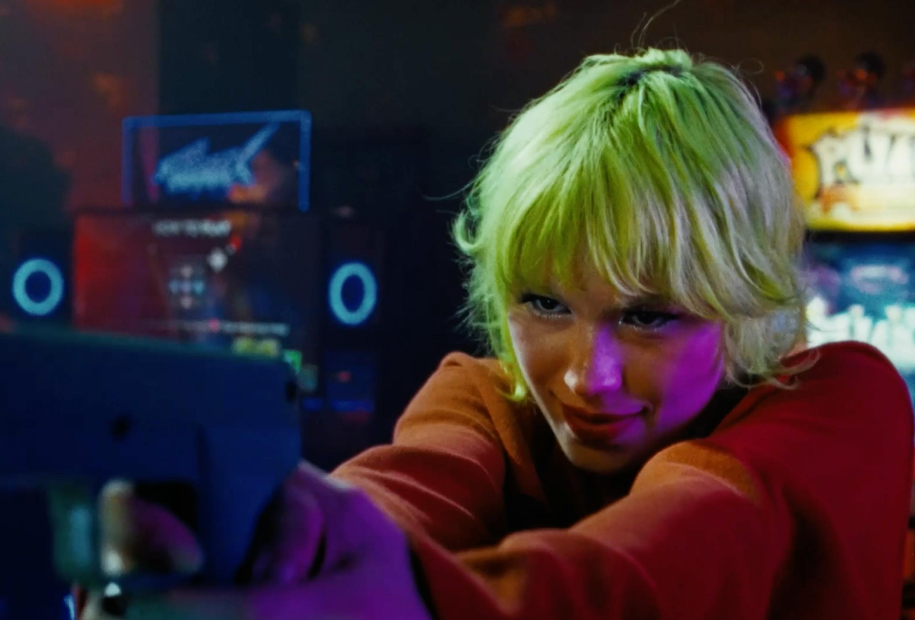 A vibrant, neon-lit shot of a woman with bright blonde hair, looking downward with colorful purple and pink light reflecting on her face.