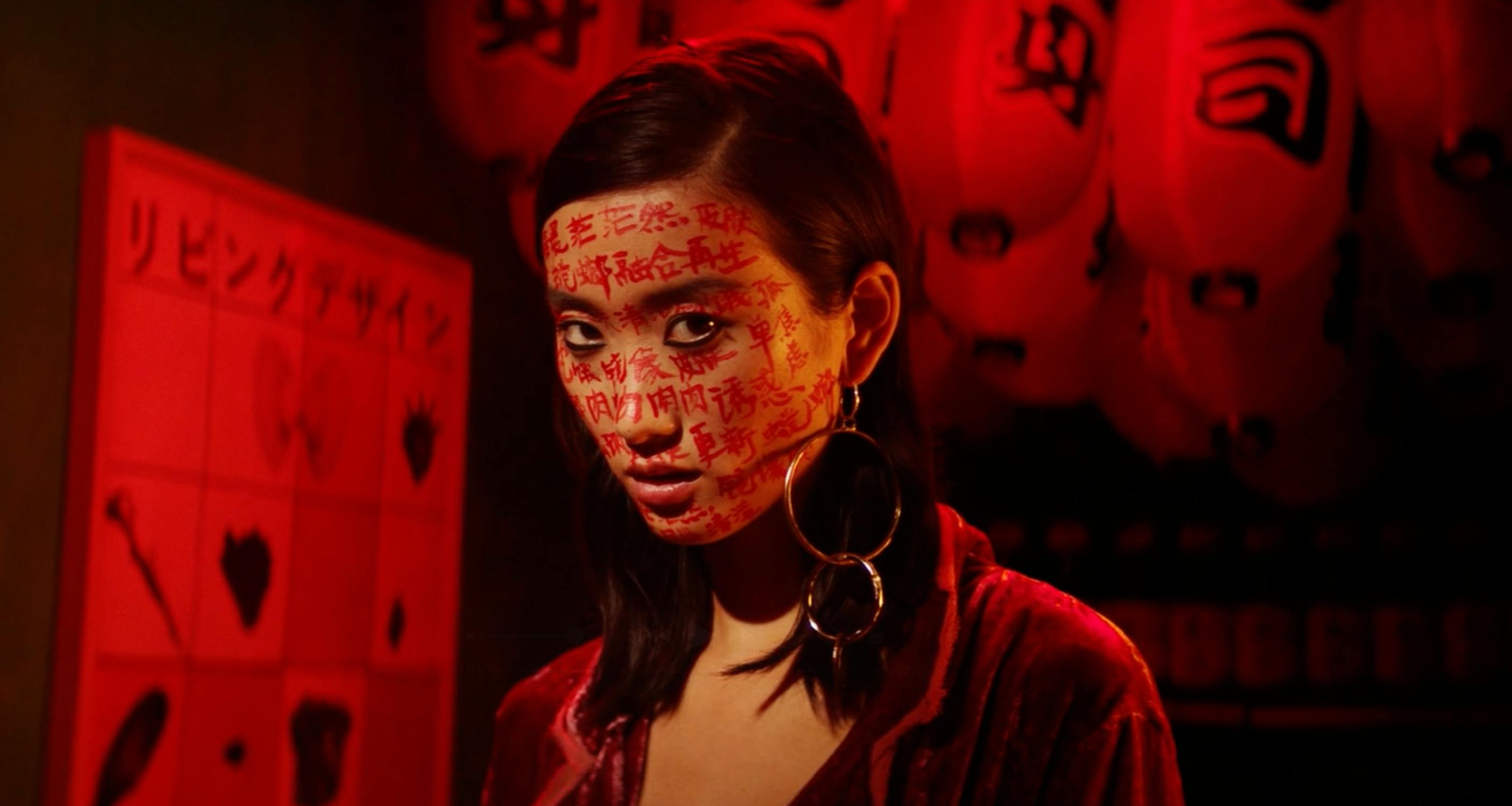 A woman in a red-lit environment looking intensely at the camera.