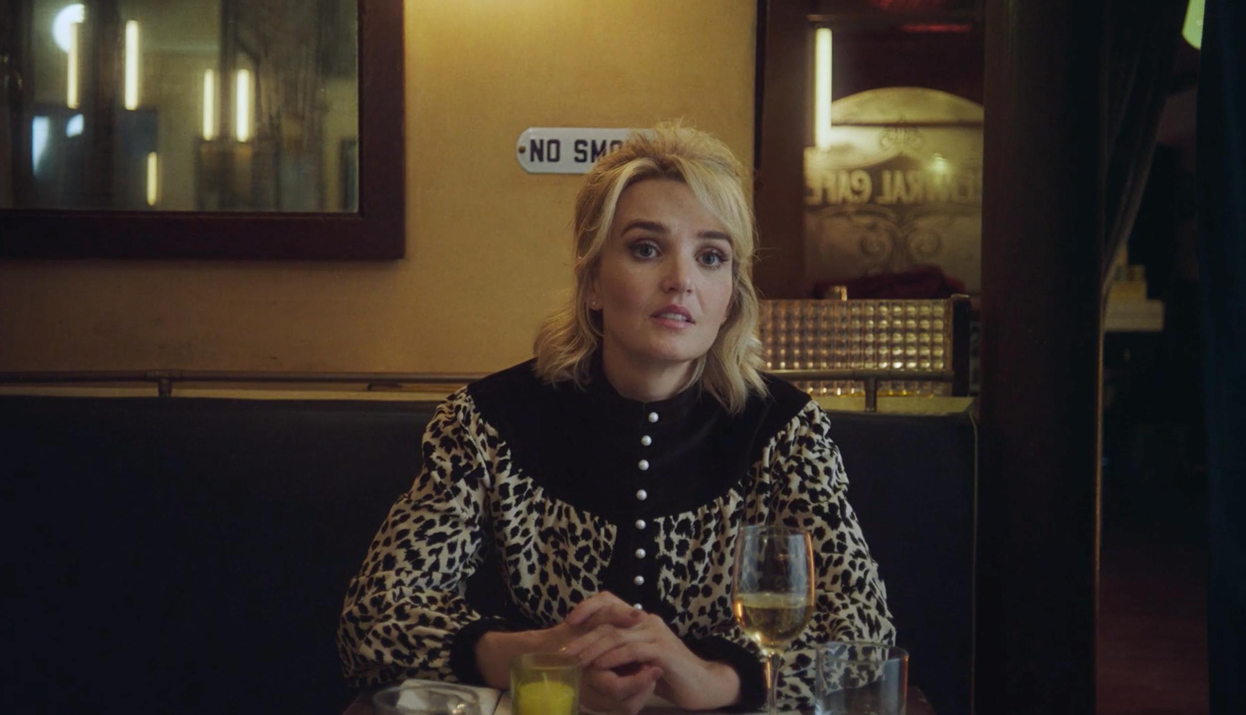 A medium shot of a woman with blonde hair and a patterned black-and-white shirt sitting in a dimly lit bar or restaurant. A classic 