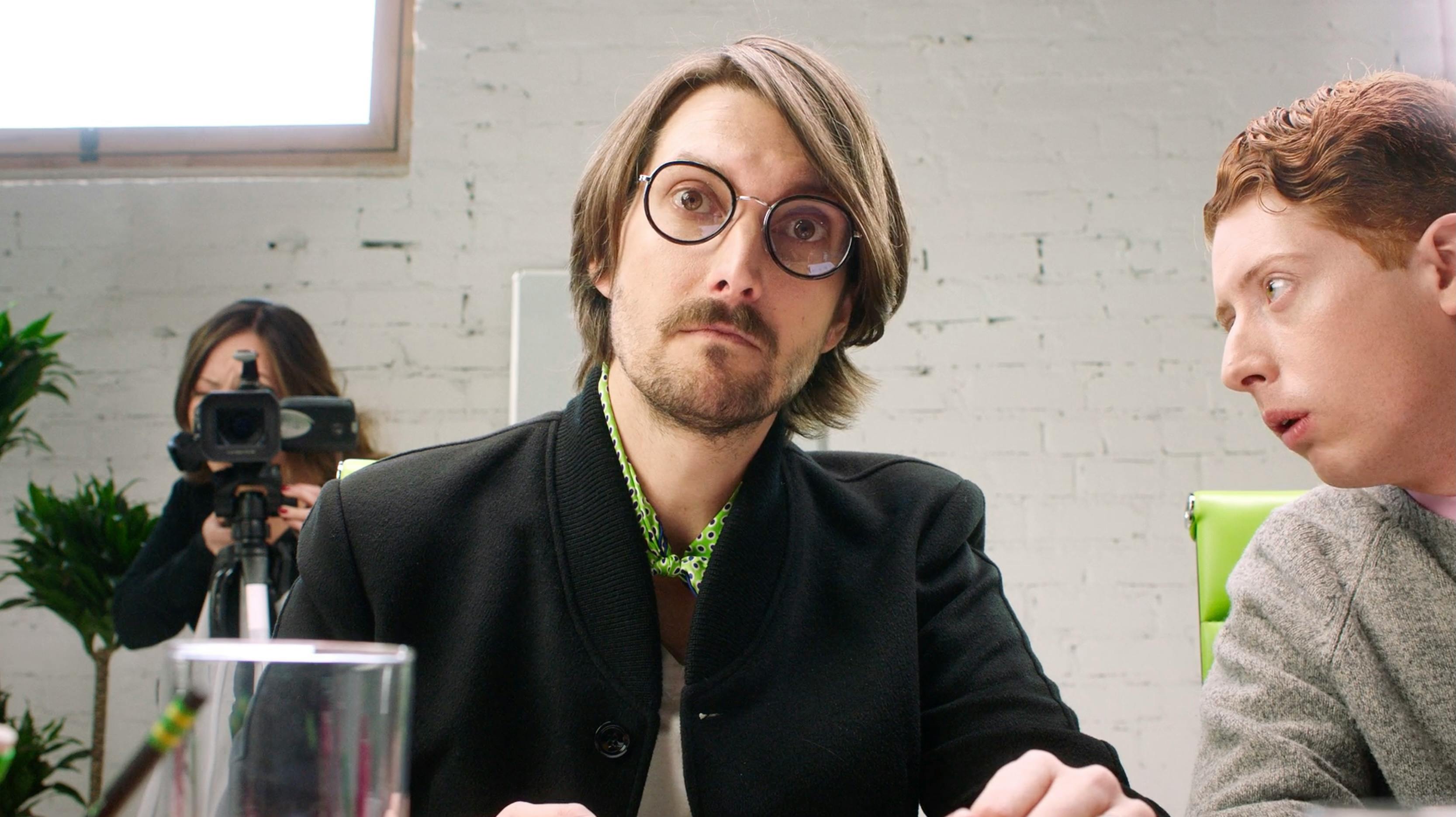 A man with glasses and a mustache looking directly at the camera in a professional setting.