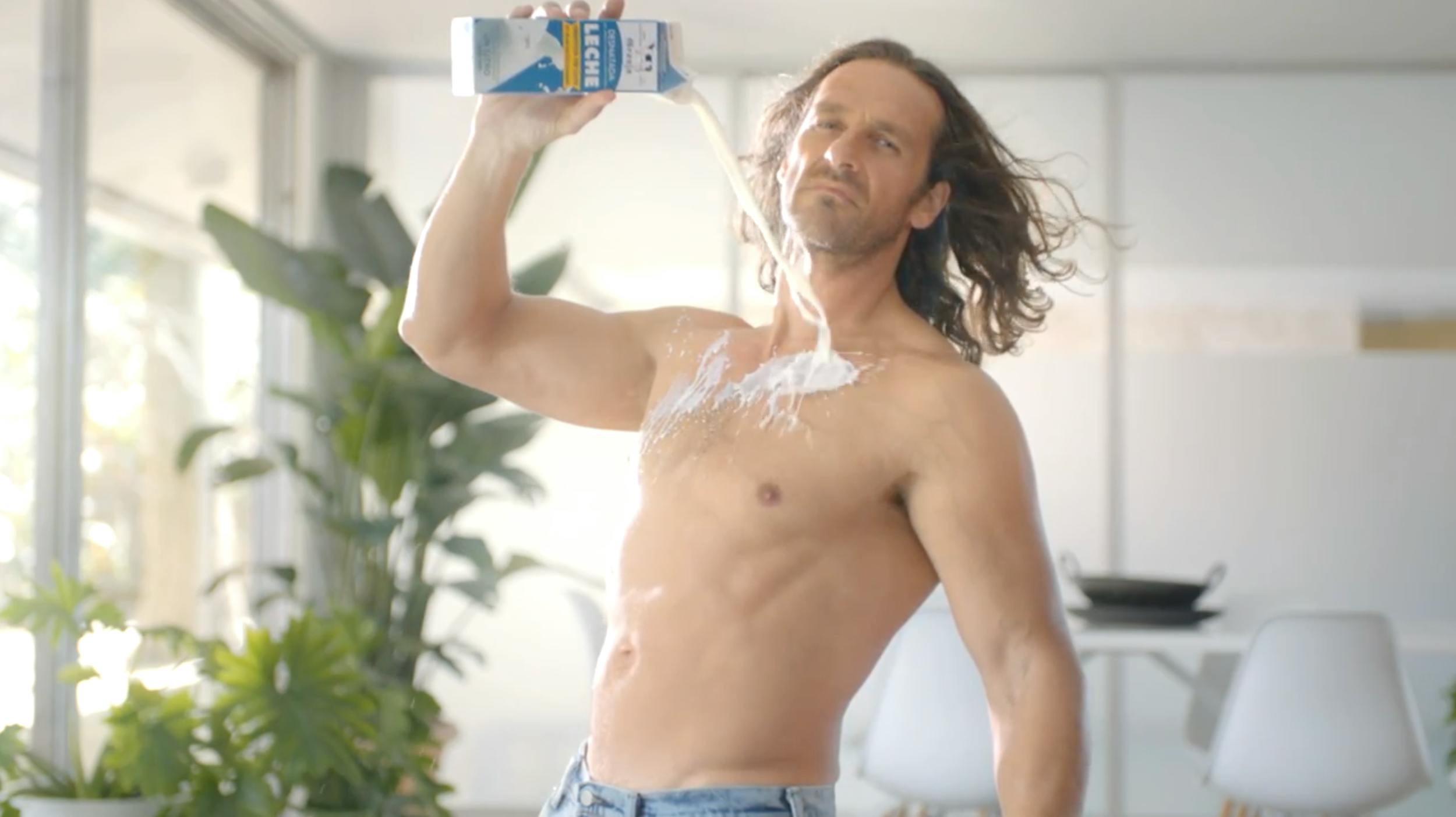A medium shot of a muscular man with long hair, shirtless in a bright, modern interior, pouring a white liquid (milk or lotion) from a carton over his chest. High-key lighting with a clean, lifestyle advertising aesthetic.