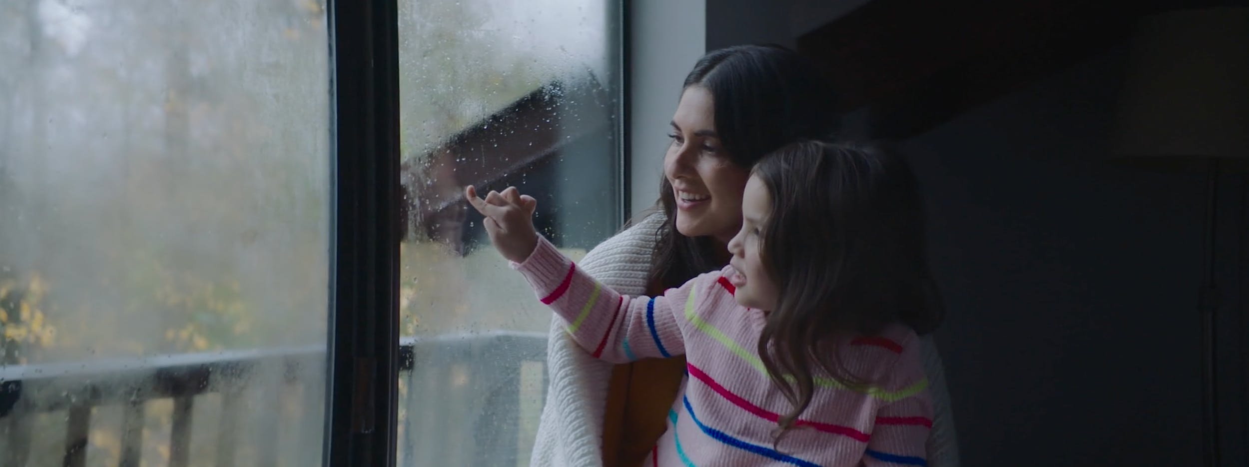 Cinematographic medium shot of mother and daughter by a wet windowpane on a film set, featuring soft key lighting and shallow depth of field, used in video production to convey warm family emotion.