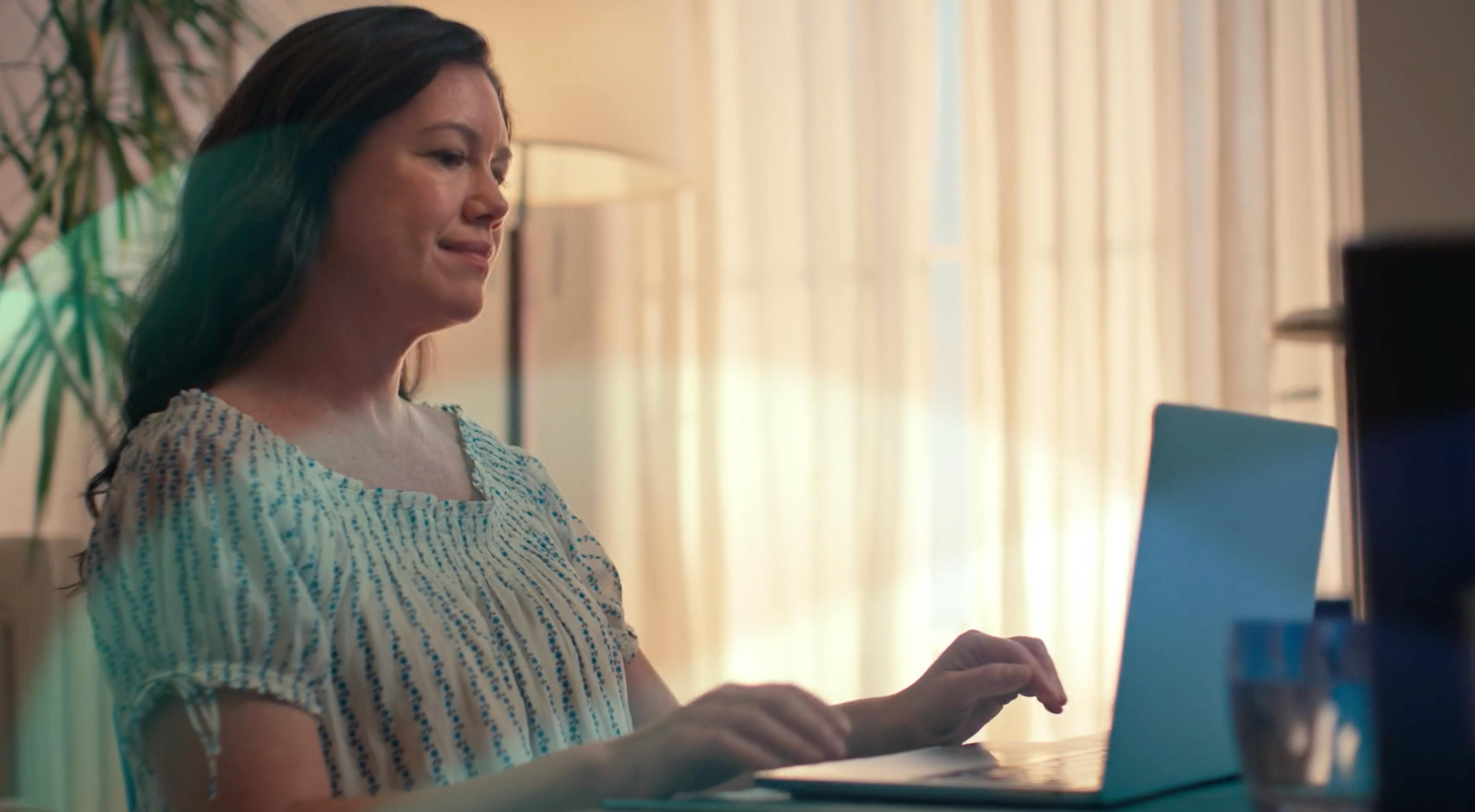 A woman sitting in a sunlit room, looking calmly toward a window.