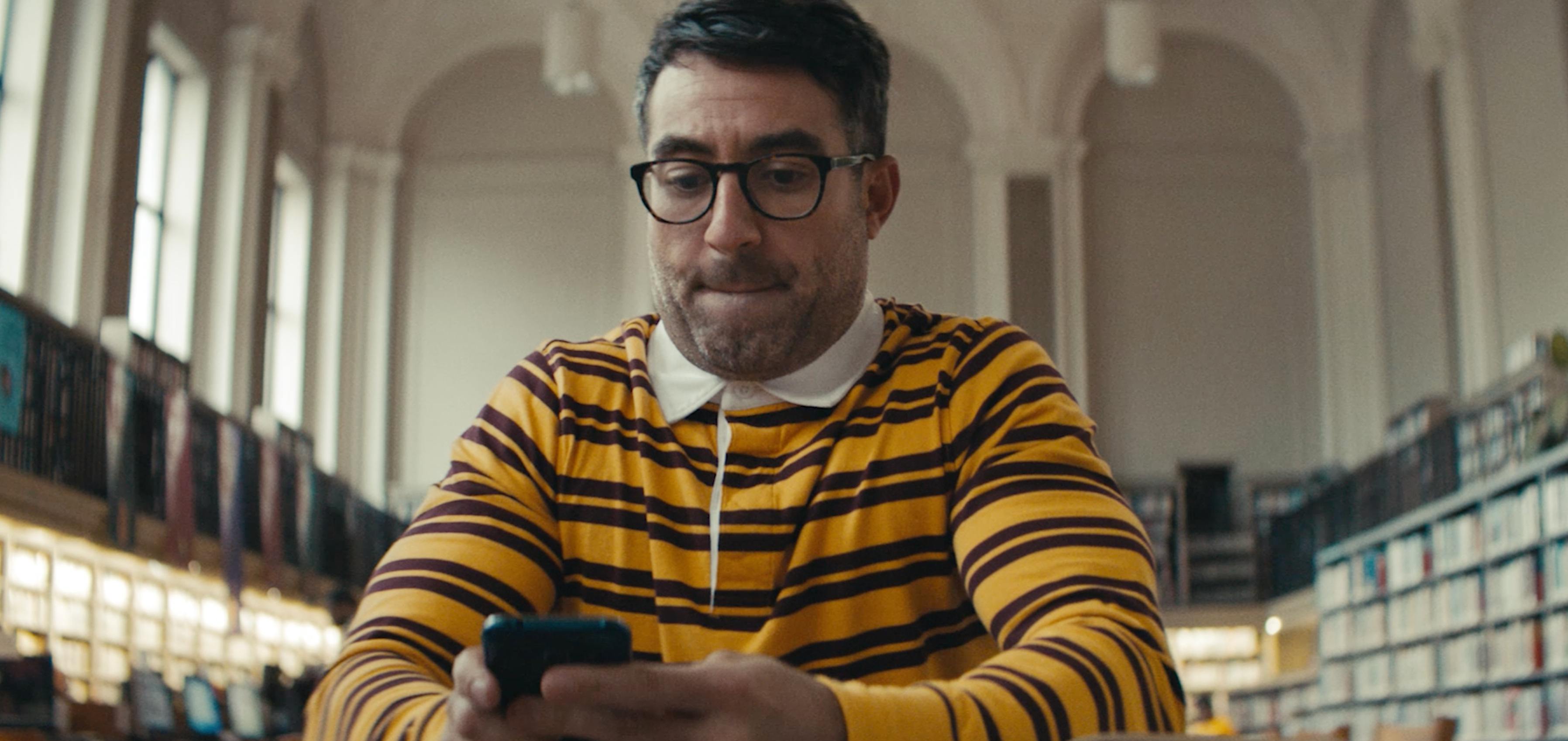 A man with glasses and a mustache, wearing a yellow striped sweater, looks down at a phone in a brightly lit indoor space.