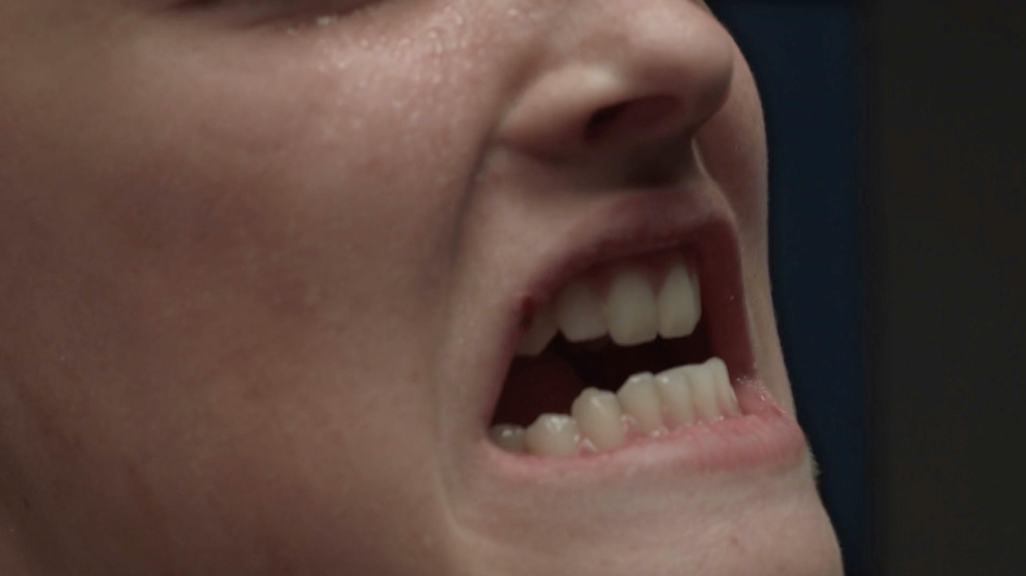 An extreme close-up of a person’s mouth, showing teeth and lips slightly parted in a grimace or expression of tension. High-detail macro photography focusing on texture and raw emotion.