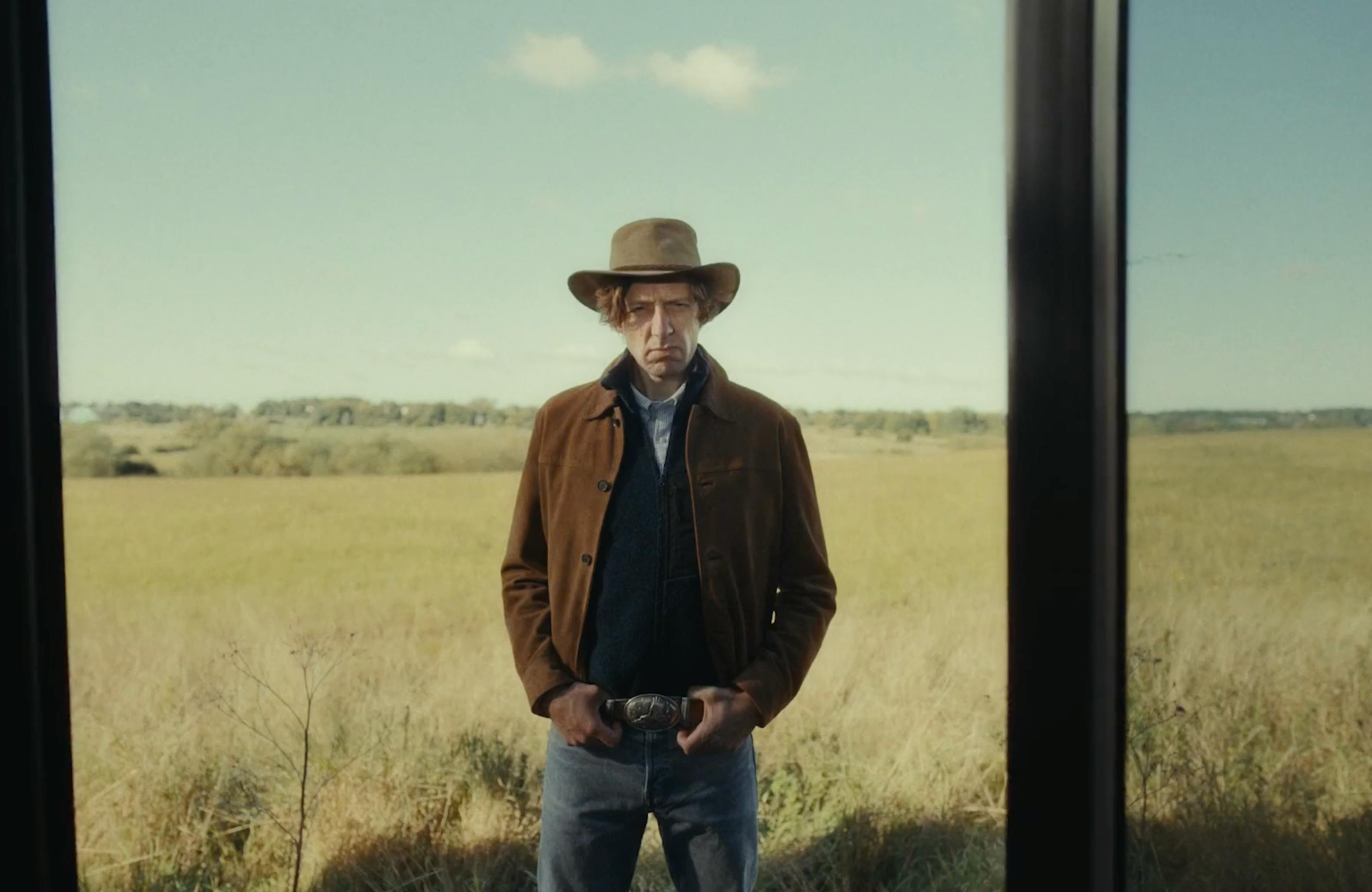 A man wearing a cowboy hat and rugged outdoor clothing stands in a vast, flat grassy field under a clear sky.